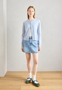 Ljusblå kofta med knappar, vit topp, denimkjol och gröna skor med blå accenter. Mot en neutral bakgrund.