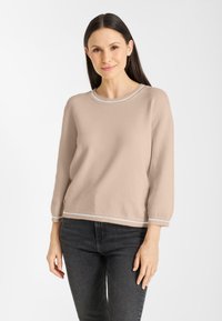 Vrouw met lang donker haar, gekleed in een beige trui met lange mouwen en witte rand, gedragen met zwarte jeans, staand tegen een witte achtergrond.