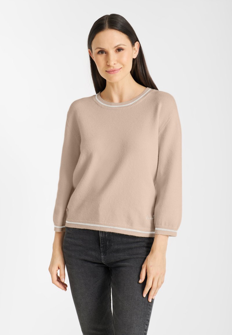 Vrouw met lang donker haar, gekleed in een beige trui met lange mouwen en witte rand, gedragen met zwarte jeans, staand tegen een witte achtergrond.