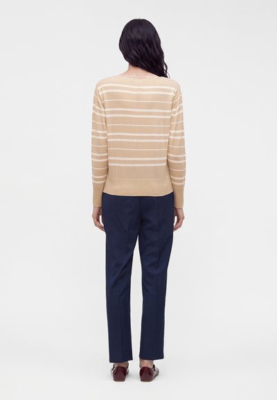 Femme aux longs cheveux noirs de dos, portant un pull rayé beige et blanc, un pantalon bleu marine et des chaussures rouge foncé.