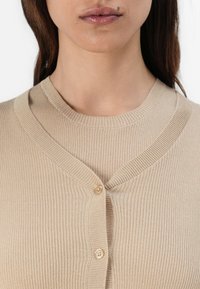 Beige gerippter Cardigan mit V-Ausschnitt, der über vier Knöpfe verfügt, aus strukturiertem Stoff gefertigt ist und eine taillierte Form hat, die den Oberkörper umschmeichelt.
