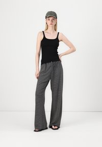 Juodas tank topas, laisvi juodai balti dryžuoti kelnes su raišteliu ties juosmeniu. Modelis dėvi pilką kepurę ir juodas šlepetes. Neutralus fonas.