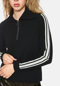 Schwarzer Zip-Up-Pullover mit geripptem Kragen, der weiße vertikale Streifen an den Ärmeln und eine strukturierte Stoffoberfläche aufweist. Nahaufnahme des Oberkörpers.
