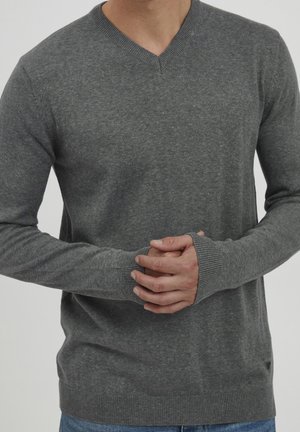 Homme portant un pull gris à manches longues et col en V, les mains jointes au niveau de la taille, sur un fond clair uni.