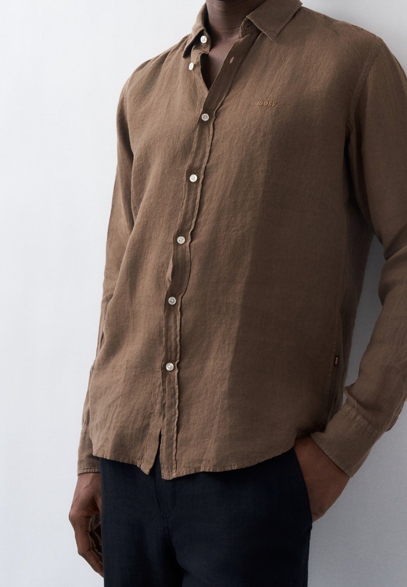 Homme portant une chemise marron à manches longues boutonnée, portée sur un pantalon foncé, une main dans la poche, sur un fond clair uni.