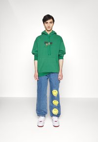 Felpa con cappuccio verde con grafica frontale, abbinata a pantaloni in denim azzurro chiaro con design di faccine sorridenti gialle. Sneakers bianche.