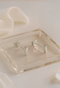 Silverfärgade studörhängen med en cylindrisk design, prydda med ingraverade mönster i varje ände, visade på en transparent bricka med mjukt tyg.