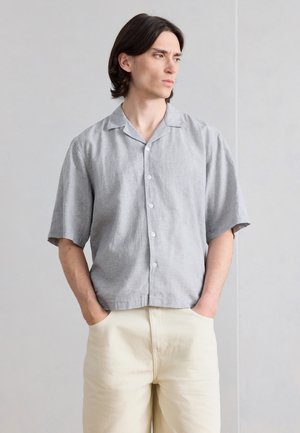 Άνδρας με σκούρα μαλλιά, που φοράει ανοιχτό γκρι κοντομάνικο πουκάμισο με κουμπιά και κρεμ παντελόνι, στέκεται με τα χέρια στις τσέπες και κοιτάζει στο πλάι.