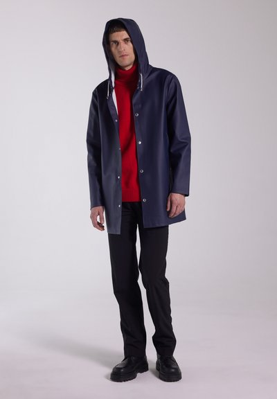 Navy waterproof coat with a hood, silver snap buttons, and a relaxed fit. Worn over a red turtleneck and paired with black pants and shoes.