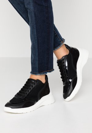 Personne portant des baskets noires avec des semelles épaisses blanches et un jean bleu foncé aux ourlets effilochés, debout sur un sol blanc.