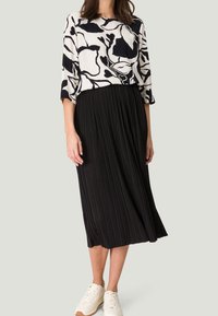 Jupe plissée noire associée à un haut blanc avec des motifs floraux abstraits noirs. Le modèle porte des baskets blanches.