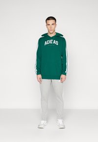 Grön hoodie med vit Adidas-logotyp, med en framstående kenguruficka och dragsnodd i huvan. Kombinerad med grå sweatpants och vita sneakers.