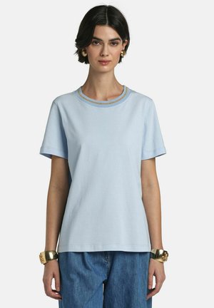 Kvinde med kort mørkt hår iført en lyseblå t-shirt med korte ærmer og guldkant ved kraven samt blå jeans, tilbehør med guldbøjler og øreringe.