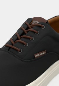 Sort sko med en tekstureret overdel, brune snørebånd og et brunt mærke med "JACK & JONES." Lys farvet sål med syning.