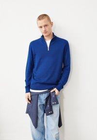 Pull à col zippé bleu en tissu tricoté, featuring un ourlet côtelé, associé à un jean délavé clair et une chemise rayée nouée à la taille.