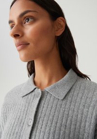 Femme à la peau marron et aux cheveux foncés portant un cardigan en tricot côtelé gris clair avec un col et des boutons, regardant sur le côté.