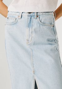 Persona che indossa una gonna di jeans azzurro chiaro con spacco frontale e una camicia bianca infilata, mani sui fianchi.
