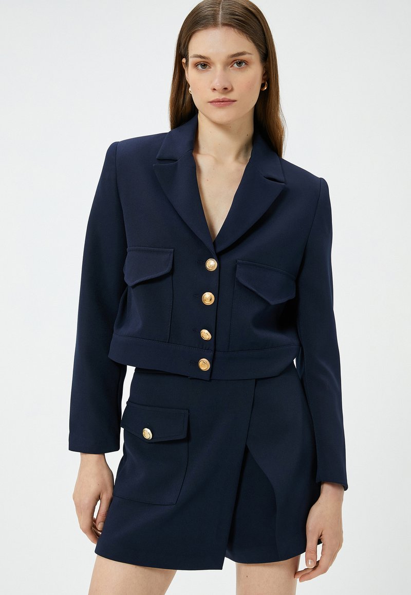 Marineblazer mit goldenen Knöpfen, mit einem cropped Design und zwei Brusttaschen. Getragen mit einem passenden hoch taillierten Rock. Glatte Textur.
