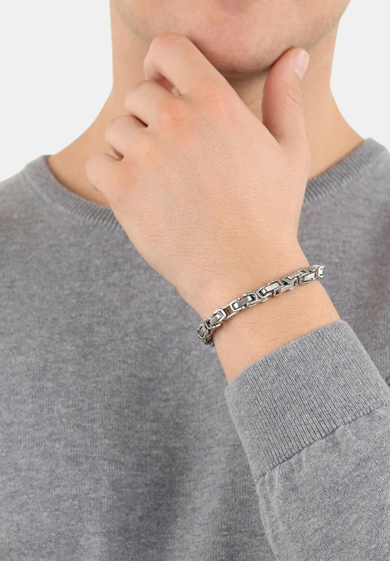 Braccialetto in metallo argento con anelli intrecciati, che presenta una finitura lucida e una texture liscia, indossato su una mano con un maglione grigio.