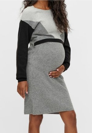 Pregnant woman wearing a grey and black color-block knit dress, gently holding her belly with one hand.