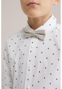 Fehér, apró geometriai mintás gombos ingben és bézs csokornyakkendőben lévő fiú, nyaktól a mellkasig látható.