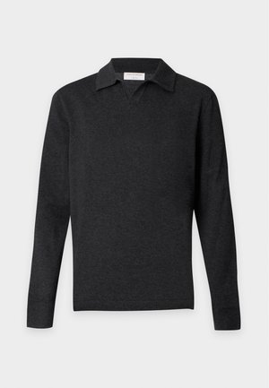 Homme debout portant un pull polo noir à manches longues, un jean bleu et des chaussures marron devant un fond clair uni.