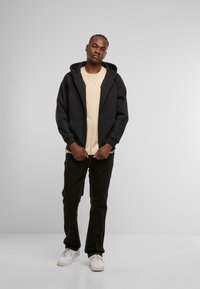Sudadera con capucha negra y bolsillos frontales, usada sobre una camiseta de color beige claro, combinada con pantalones negros y zapatillas blancas. Textura de tela suave.