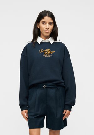 Femme aux longs cheveux foncés portant un sweat-shirt Tommy Hilfiger bleu marine sur une chemise blanche à col et un short bleu marine ceinturé, regardant vers l'avant.