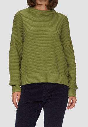 Kvinde iført en løstsiddende ribbet olivengrøn sweater med lange ærmer og mørkeblå bukser mod en ensfarvet baggrund.