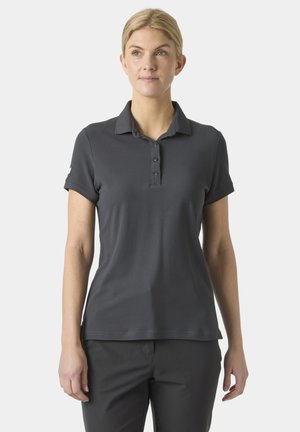 Žena v tmavě šedé krátkosrsté polokošili a odpovídajících kalhotách stojí na jednobarevném světlém pozadí.