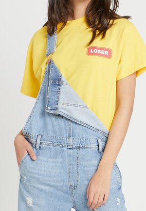 Keltainen t-paita, jossa on "LOSER"-teksti, yhdistetty vaaleansinisiin farkkuhaalareihin, joissa on kulutuspintainen tekstiili, säädettävät olkaimet ja etutaskut.