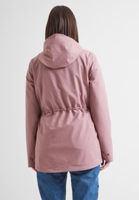Person wearing a pink hooded jacket with cinched waist and blue jeans, facing away against a plain light background.