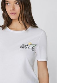 Weißes Baumwoll-T-Shirt mit kurzen Ärmeln, verziert mit schwarzer und grüner Stickerei eines Margarita-Glases und dem Satz "Ich brauche einen MARGARITA."