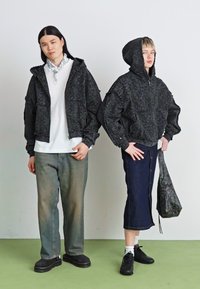 Two individuals wear dark textured jackets with hoods. One has a white shirt and faded jeans; the other wears a denim skirt and handbag.