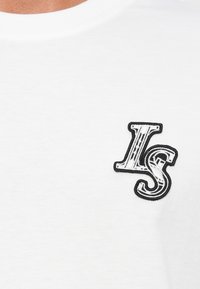Hvitt bomulls T-skjorte med en svart brodert "LS" logo på venstre bryst, rund halslinning og glatt tekstur.