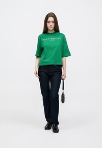 Πράσινο βαμβακερό μπλουζάκι με στρογγυλή λαιμόκοψη και κοντά μανίκια, με λευκό κείμενο "TOMMY HILFIGER EST. 1985". Συνδυασμένο με σκούρο μπλε τζιν και μαύρα παπούτσια.