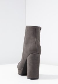 Harmaan tekonahkainen nilkkabootsi, jossa on korkea loiva korko, pyöreä varvas ja sivuvetoketju. Sileä pinta, jossa on minimaalinen ommelkoristelu.