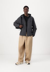 Donkergrijze hooded jack met rits aan de voorkant, beige relaxed-fit broek en beige sneakers. Model houdt een zwarte rugtasstrap vast.