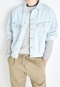 Light blue denim jacket with silver hardware, front pockets, and yellow stitching, layered over a grey ribbed sweater and beige pants.