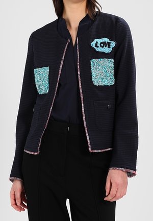 Femme portant une veste noire texturée avec des poches en sequins bleus et un patch "LOVE" sur la poitrine, par-dessus un chemisier et un pantalon noirs.