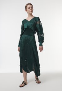 Largo vestido plisado en verde oscuro con un corpiño entallado, escote en V y acentos de mangas de encaje transparentes, combinado con sandalias negras con herrajes dorados.