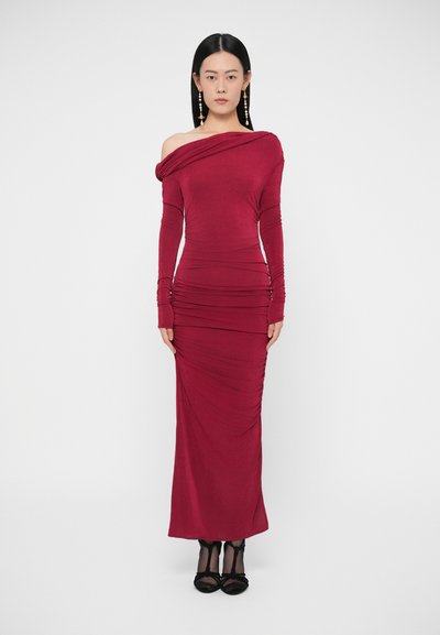 Mujer de pie con un vestido largo burdeos ajustado y con los hombros descubiertos, con detalles fruncidos, y zapatos de tacón negros, sobre un fondo liso.