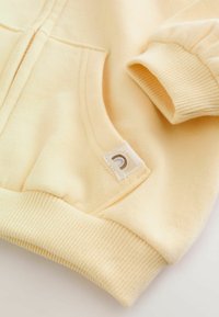 Gele hoodie met een kangoeroezak, geribbelde manchetten en een klein label met een gebogen ontwerp nabij de onderzoom. Zachte stoftextuur.