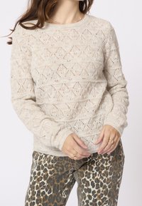 Femme portant un pull en maille crème avec un motif en losange et un pantalon à imprimé léopard, montrant des bagues en or aux doigts.