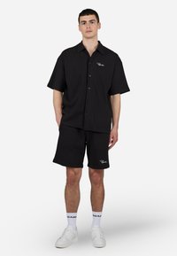 Cămașă neagră cu mâneci scurte, cu nasturi, având un model texturat și broderie cu logo, asortată cu pantaloni scurți negri și adidași albi.