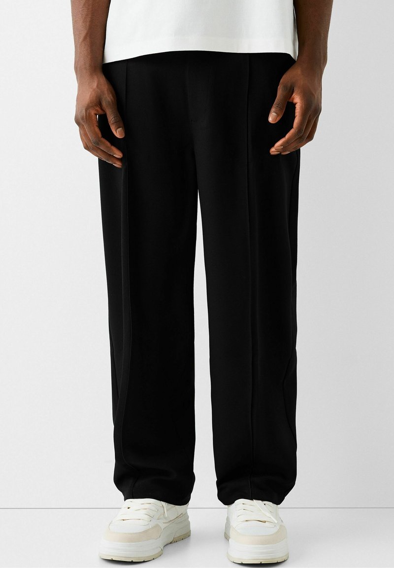 Personne portant un pantalon noir ample et des baskets blanches, debout devant un fond gris clair uni.