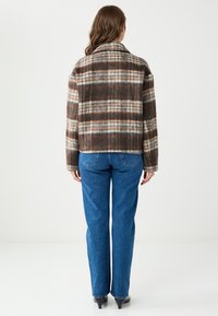 Veste en laine à carreaux marron avec un col à revers, une coupe carrée et une fermeture à boutons. Associée à un jean bleu à jambes droites.