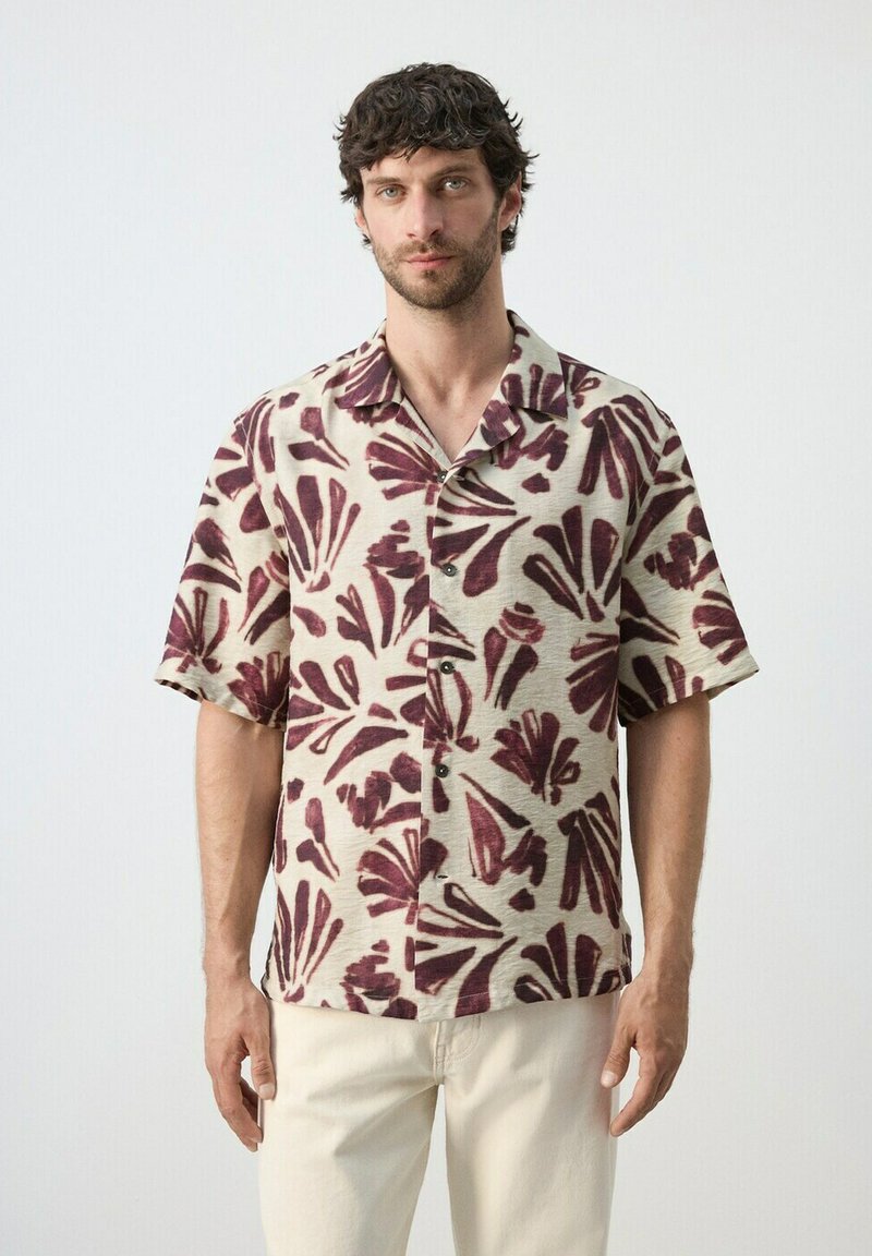Homme portant une chemise beige à manches courtes avec un motif floral abstrait rouge foncé et un pantalon de couleur claire, debout devant un fond uni.