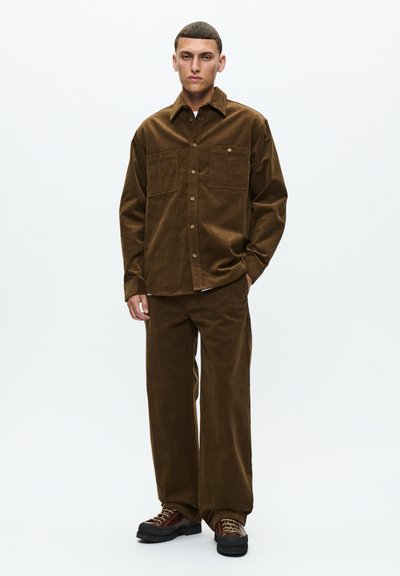 Jeune homme portant une chemise et un pantalon en velours côtelé brun avec des chaussures à lacets marron, debout sur un fond blanc.