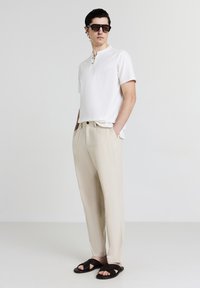 Uomo che indossa una maglietta henley a maniche corte bianca, pantaloni beige, sandali neri intrecciati e occhiali da sole scuri, in piedi con le mani in tasca.